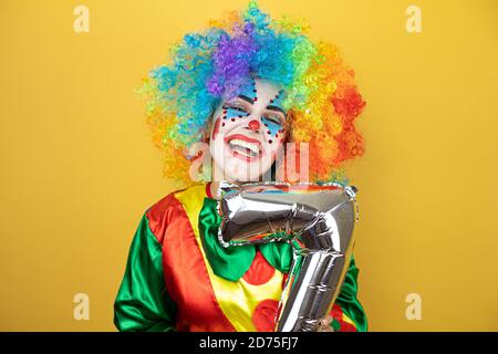 Clown se tenant sur fond jaune insolé et tenant un ballon numéro sept. Anniversaire Banque D'Images