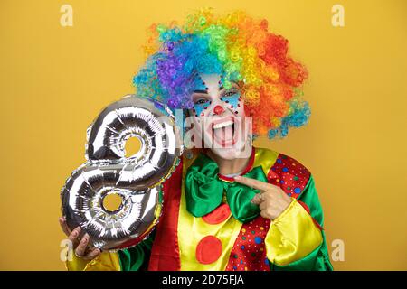 Clown se tenant sur fond jaune insolé et tenant un ballon numéro huit. Anniversaire Banque D'Images