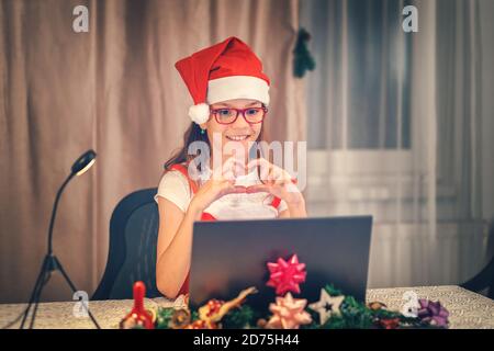 Bonne petite fille qui fait le cœur avec les mains sur appel vidéo avec les parents et la famille célébrant noël en ligne Banque D'Images