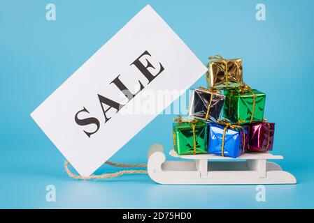 Joyeux noël ! Photo de mini-traîneaux de jouet transportant lot de grande pile énorme de mini petites boîtes présentes avec affiche de vente isolée sur bleu Banque D'Images