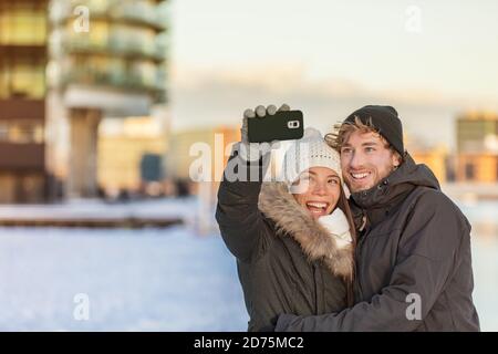 Selfie couple heureux hiver voyage marcher touristes prenant photo avec le téléphone sur la ville rue panorama style de vie. Femme asiatique, homme caucasien portant une maille Banque D'Images