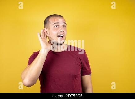 Homme russe portant un t-shirt rouge de base sur fond jaune insolé souriant avec la main sur l'oreille écoutant une audition à la rumeur ou des potins Banque D'Images