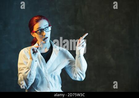 Infirmière ou médecin sur fond sombre avec stylo à la main. Femme en lunettes et uniforme médical blanc pointe sur le côté. Concept de soins de santé avec espace de copie. Banque D'Images