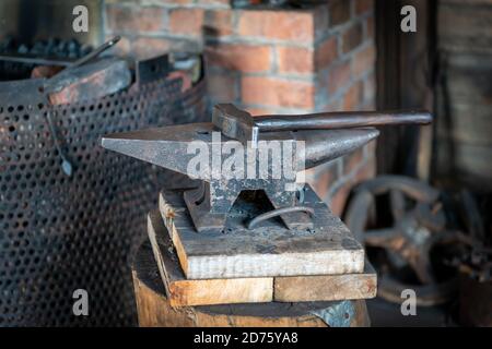 Une enclume vintage et un marteau sur des blocs de bois dans une forge ou un atelier. Il y a un foyer en acier dans le fond rempli de charbon. Banque D'Images