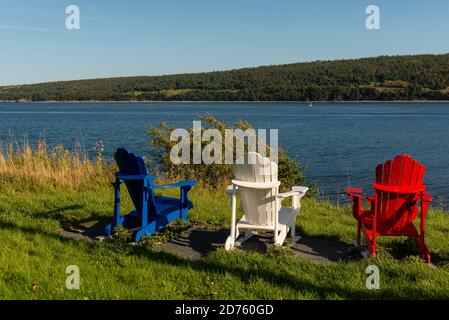 Trois chaises adirondack un rouge, bleu et blanc se trouve sur le bord de l'océan pendant une journée ensoleillée d'été. Il y a une montagne couverte d'arbres au loin. Banque D'Images