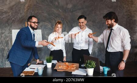 Des collègues heureux et variés passent une pause de travail ensemble en mangeant une pizza à emporter au bureau, les employés multiraciaux souriants s'amusent, rient et bavarent Banque D'Images