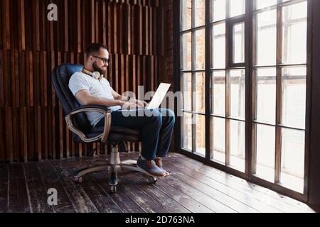 Un homme d'affaires indépendant dans un t-shirt blanc et des lunettes travaille sur un ordinateur portable. Tapez le texte sur le clavier ordinateur assis dans une chaise dans un bureau confortable Banque D'Images