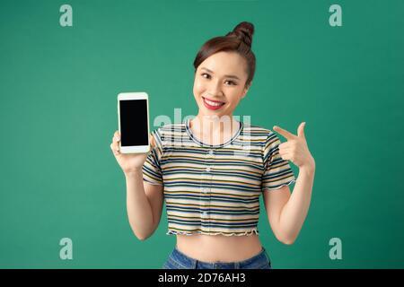 Jeune femme asiatique souriante pointant avec un index sur un téléphone à écran vierge isolé sur fond vert. Banque D'Images