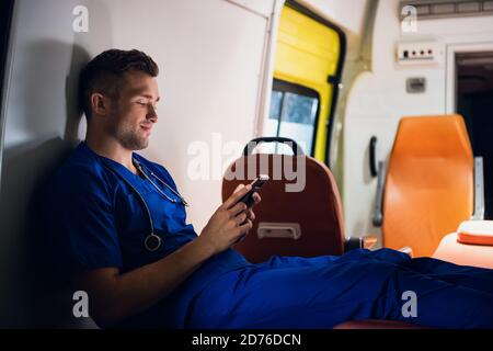 Jeune médecin dans un uniforme bleu se reposant et discutant avec quelqu'un au téléphone la nuit. Banque D'Images