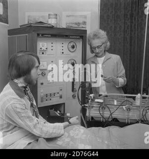 Années 1970, hôpital, un homme paent assis sur un lit d'hôpital ayant un traitement de dialyse, via une unité Cambridge Diaysis Mark 5 machine, adminée par une femme technicien au chevet, Lewisham, sud-est de Londres, Angleterre, Royaume-Uni. Un tel traitement est utilisé lorsque les organes vitaux du corps ne fonctionnent pas correctement. Ici l'homme a la dialyse pour ses reins qui ne fonctionnent pas comme ils devraient. La première dialyse réussie a eu lieu en 1943. Banque D'Images