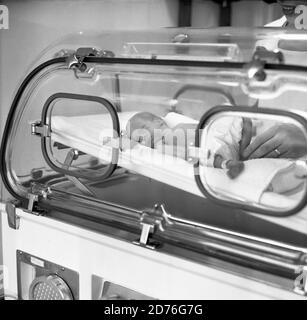 années 1970, historique, un nouveau-né prématuré bébé dans un incubateur. On peut voir à quel point le bébé prématuré est minuscule en comparant la taille de ses pieds de jambes avec les doigts de la main de l'infirmière vu dans l'image sur la droite. Un incubateur est un équipement médical qui protège la préemie contre les infections et les allergènes éventuels ou le bruit excessif des niveaux de lumière qui pourraient la causer. En outre, ils sont nourris dans des incubateurs pour les maintenir au chaud pendant que l'humidité de l'air est régulée de sorte qu'il s'agit d'un environnement contrôlé et protecteur pour leurs soins. Banque D'Images