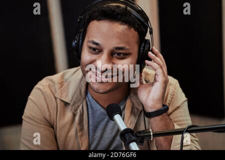 Gros plan sur un jeune hôte de radio homme heureux souriant de côté pendant la diffusion en studio, à l'aide d'un microphone et d'un casque Banque D'Images