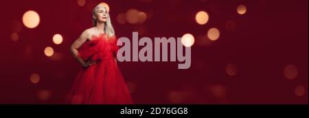 Belle femme âgée dans une robe rouge qui regarde loin et sourire. Belle femme mûre posant sur fond rouge. Banque D'Images