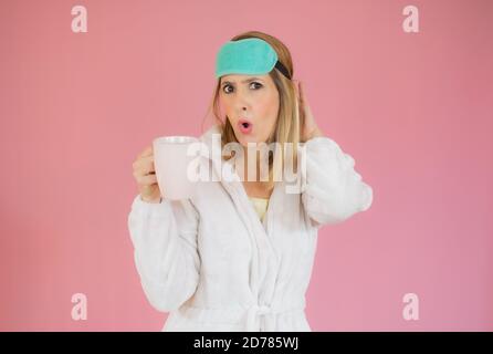 Jeune femme portant un pyjama et un masque de sommeil buvant du café sur fond rose isolé. Banque D'Images