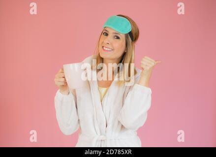 Jeune femme portant un pyjama et un masque de sommeil buvant du café sur fond rose isolé. Banque D'Images