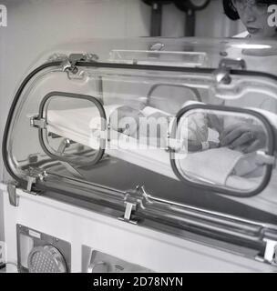 années 1970, historique, un nouveau-né prématuré bébé dans un incubateur. On peut voir à quel point le bébé prématuré est minuscule en comparant la taille de ses pieds de jambes avec les doigts de la main de l'infirmière vu dans l'image sur la droite. Un incubateur est un équipement médical qui protège la préemie contre les infections et les allergènes éventuels ou le bruit excessif des niveaux de lumière qui pourraient la causer. En outre, ils sont nourris dans des incubateurs pour les maintenir au chaud pendant que l'humidité de l'air est régulée de sorte qu'il s'agit d'un environnement contrôlé et protecteur pour leurs soins. Banque D'Images