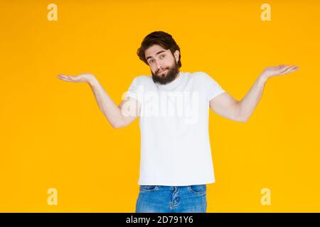 Un jeune homme avec une barbe dans un T-shirt blanc écartant ses bras, choqué par l'expression inattendue, hésite à dire, la réponse n'a aucune idée. JE Banque D'Images