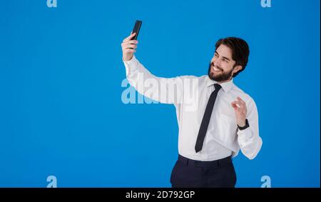 Jeune homme d'affaires barbu parlant au téléphone sur facetime . Un homme dans une chemise blanche et cravate prend une photo de selfie sur un fond bleu Banque D'Images