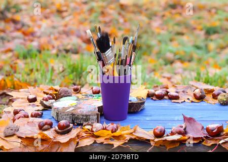Lieu de travail pour peintre dans un parc d'automne avec palette de bois et pinceaux dans un récipient en plastique. Feuilles d'automne et châtaignes sur fond de bois bleu. Banque D'Images