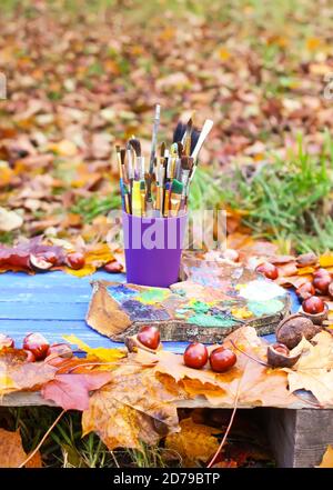 Lieu de travail pour peintre dans un parc d'automne avec palette de bois et pinceaux dans un récipient en plastique. Feuilles d'automne et châtaignes sur fond de bois bleu. Banque D'Images