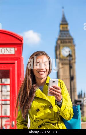 Une femme de ville asiatique envoie des SMS sur un téléphone mobile à pied dans la rue de Londres. Bonne fille chinoise en manteau de pluie jaune de printemps à l'extérieur avec fond Big Ben, Royaume-Uni. Banque D'Images