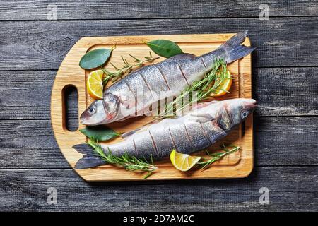 Bar de mer fruits de mer frais crus évidés et détartré sur une planche à découper avec du romarin frais, des quartiers de citron et une feuille de Laurier sur une planche à découper sur un b en bois sombre Banque D'Images