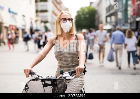 Femme à vélo dans la rue de la ville portant un masque médical en public pour prévenir la propagation du virus corona. Nouvelle normale pendant l'épidémie de covid. Banque D'Images