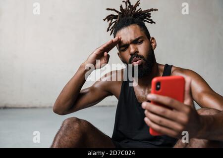 Image d'un jeune sportif afro-américain qui fait des gestes tout en utilisant le mobile téléphone à l'intérieur Banque D'Images