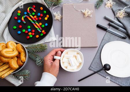 Main avec une tasse de café, des sucettes, des biscuits, des guirlandes de Noël, un bloc-notes vierge, un stylo et des branches de sapin sur fond gris. Compositi de Noël Banque D'Images
