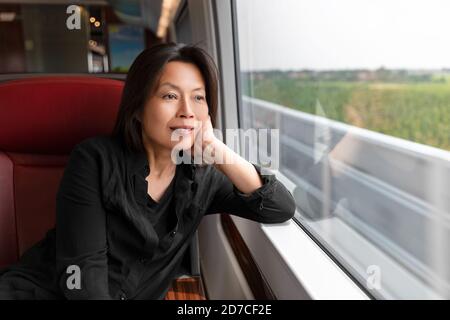 Femme asiatique d'âge moyen regardant par la fenêtre à vue sur le paysage assis sur le train Voyage style de vie. Passager chinois mûr voyageant souriant Banque D'Images