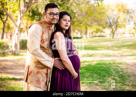 Jeune femme enceinte asiatique indienne avec son mari portant une tenue traditionnelle debout dans un parc ou un jardin pour écrire du texte. Banque D'Images