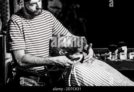 Un homme barbu assis dans un fauteuil dans un salon de coiffure, tandis qu'un coiffeur raque sa barbe avec un rasoir dangereux. Coiffeur rasant un barbu dans un Banque D'Images