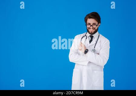 Un jeune barbu médecin sur fond bleu dans un manteau blanc regarde la caméra. COVID-19. Auto-isolation. Concept de séjour à la maison. Quarantaine. Bannière Banque D'Images