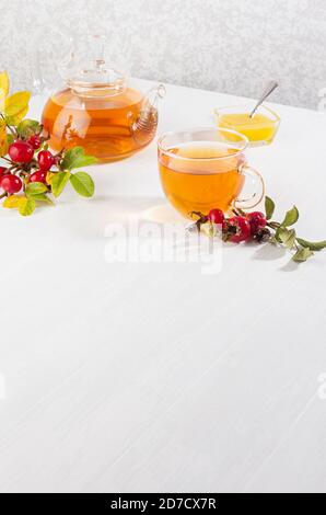 Thé d'automne confortable avec thé aux hanches roses dans une théière avec tasse, miel, branche de rose chien sur table en bois blanc clair et doux, espace de copie, verticale. Banque D'Images