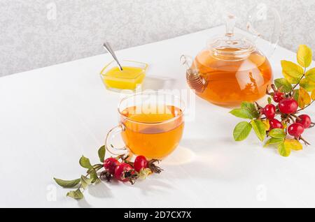 Thé d'automne confortable avec thé aux hanches roses dans une théière avec tasse, miel, branche de rose chien sur une table en bois blanc clair et doux, espace de copie. Banque D'Images