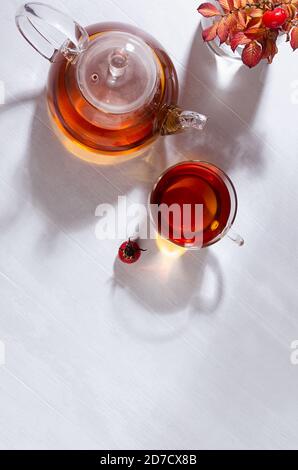 Thé d'automne ensoleillé avec thé aux hanches roses avec théière, tasse, branche orange aux hanches roses avec ombres sur la table en bois blanc, espace de copie, verticale. Banque D'Images