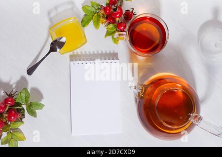 Heure de thé d'automne avec thé rosehip avec théière, tasse, branche de hanche rose d'automne et bloc-notes vierge par temps ensoleillé avec ombres sur table en bois blanc vue sur le dessus, cop Banque D'Images