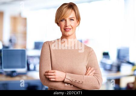 Portrait d'une belle femme d'affaires blonde debout avec les bras croisés au bureau tout en regardant l'appareil photo et en souriant. Banque D'Images