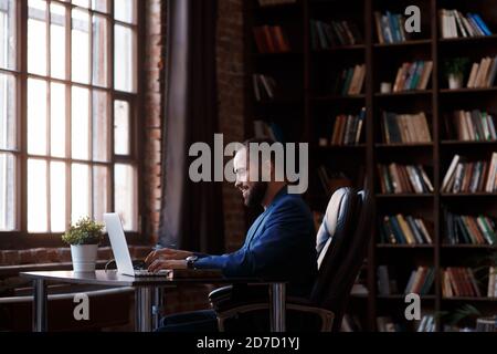 Un jeune homme d'affaires travaille sur un ordinateur portable dans un bureau luxueux de la bibliothèque. Un homme barbu, gai dans un costume bleu à un bureau est de taper sur un clavier Banque D'Images