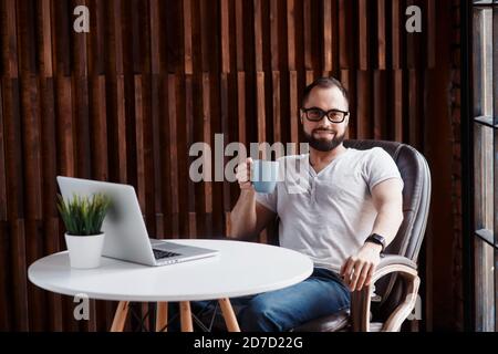 Jeune homme d'affaires barbu avec succès freelance dans un collègue de bureau. Happy man programmeur développeur dans un t-shirt blanc souriant et buvant du café à Banque D'Images
