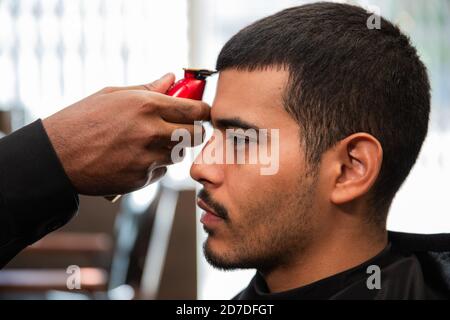 Un coiffeur afro-américain noir coupe et coupe les cheveux d'un homme latino-américain hispanique avec un bouc et une moustache avec un rasoir électrique dans un salon de coiffure. Banque D'Images