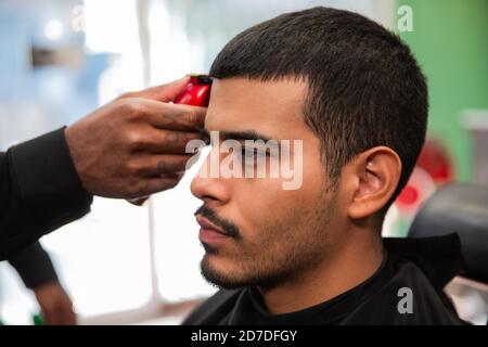 Un coiffeur afro-américain noir coupe et coupe les cheveux d'un homme latino-américain hispanique avec un bouc et une moustache avec un rasoir électrique dans un salon de coiffure. Banque D'Images