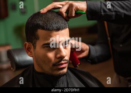 Un coiffeur afro-américain noir coupe et coupe les cheveux d'un homme latino-américain hispanique avec un bouc et une moustache avec un rasoir électrique dans un salon de coiffure. Banque D'Images