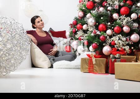 joyeux noël, femme enceinte heureuse près de l'arbre illuminé et décoré avec des paquets de cadeaux sous emballage, joyeux et souriant assis dans la liv Banque D'Images