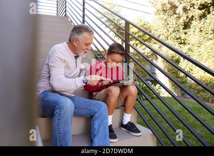 Grand-père assis sur des marches à l'extérieur à la maison avec petit-fils utilisant Tablette numérique Banque D'Images