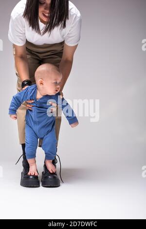 Mère encourageant bébé enfant à prendre les premiers pas. Un bébé pieds nus en costume bleu se tient sur les bottes de mather. Maman garde bébé en sécurité avec ses mains. Banque D'Images