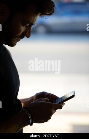 Silhouette de l'homme envoyant un message texte sur un téléphone portable en position debout près de la fenêtre Banque D'Images