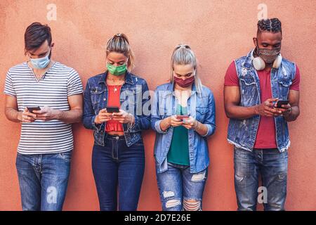 Groupe de personnes utilisant leur smartphone dans Covid 19 fois protégé avec masque de visage - amis vérifiant les nouvelles en ligne tout en se tenant près du mur et holdi Banque D'Images