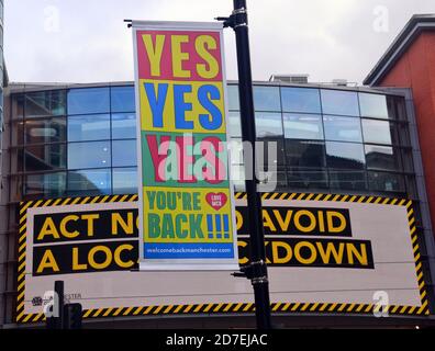 Covid 19 ou coronavirus signalisation et publicité à Manchester, Grand Manchester, Angleterre, Royaume-Uni. Une bannière sur un poteau d'éclairage dans le centre de Manchester accueille les gens de retour au centre-ville tandis que derrière lui un panneau lumineux payé par le conseil municipal de Manchester sur le centre commercial Arndale invite les gens à agir maintenant pour éviter un verrouillage local. Banque D'Images