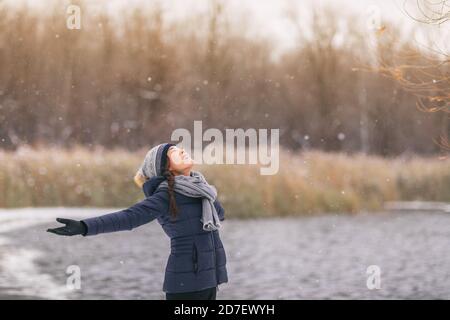 Hiver bonne fille appréciant neige neige neige chute flocons de neige tombant. Femme asiatique aux bras ouverts portant une écharpe temps froid, un chapeau, des gants veste chaude marche Banque D'Images